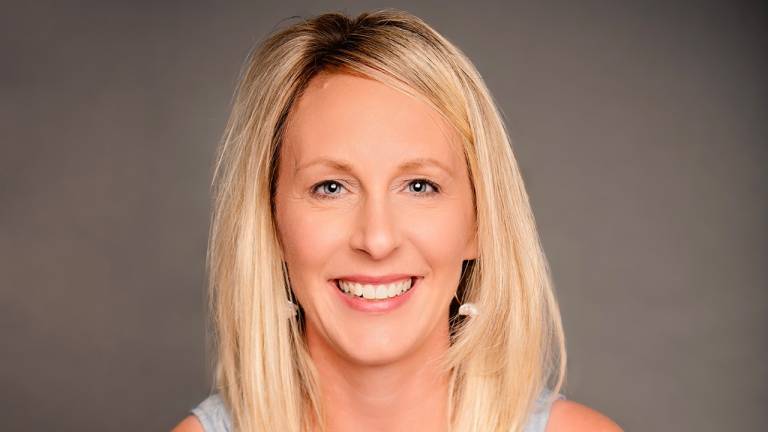 Portrait of a Jennifer Mitchell with blonde hair, wearing earrings, against a neutral gray background.