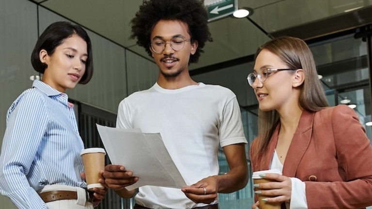 Three colleagues standing together, holding a document and coffee cups, having a discussion.