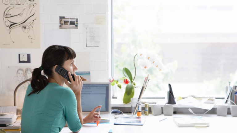 Woman talking on the phone while sitting at a desk with a computer in a brightly lit room.