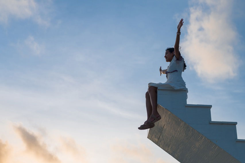 Asian woman raise arms up sit on top of wooden staircase with sunset sky in the background, victory, success concept
